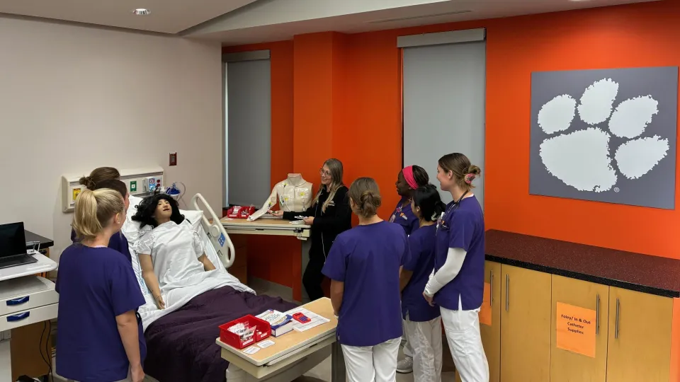 One professor in black and six students in purple and white scrubs in a nursing clinical lab.